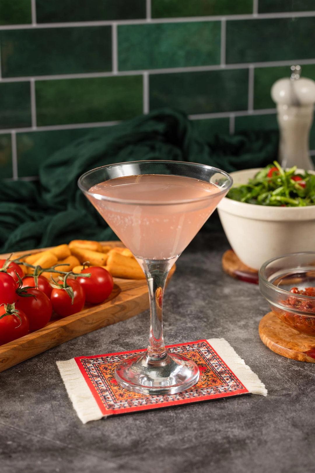Cocktail in a martini glass on a coaster with tomatoes and bread in the background.