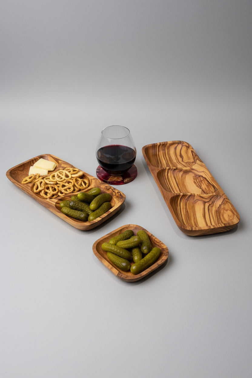 A wooden divided serving tray with three compartments, styled on a light wood table alongside a glass of red wine, pretzels, and pickles.