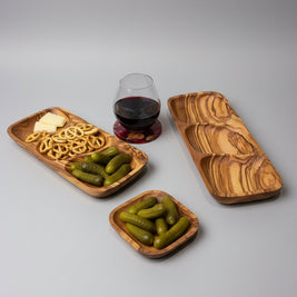 A wooden divided serving tray with three compartments, styled on a light wood table alongside a glass of red wine, pretzels, and pickles.