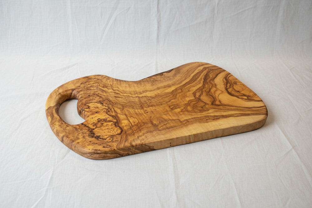 A rustic olive wood serving board with a natural live edge and a cutout handle, displayed on a light wood floor.