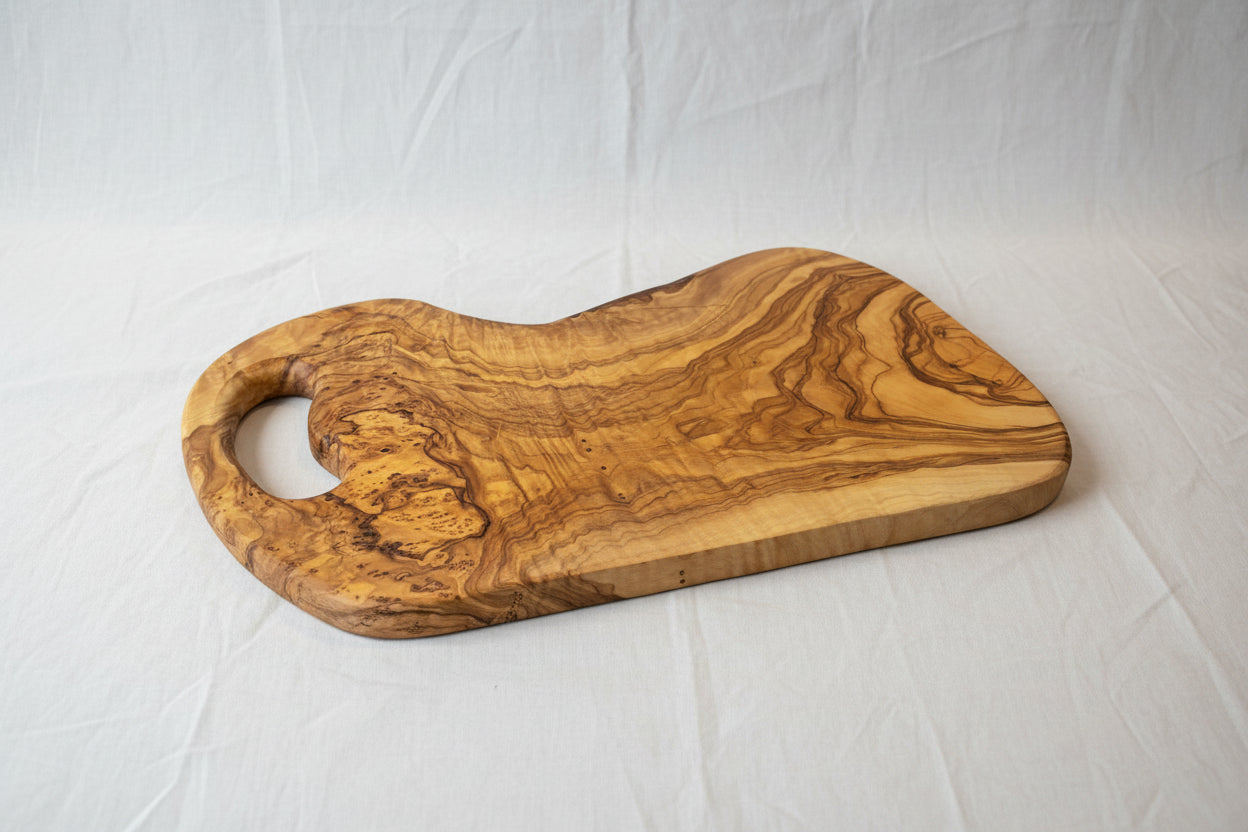 A rustic olive wood serving board with a natural live edge and a cutout handle, displayed on a light wood floor.
