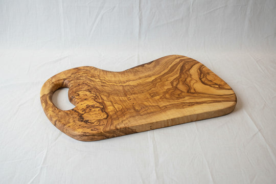 A rustic olive wood serving board with a natural live edge and a cutout handle, displayed on a light wood floor.