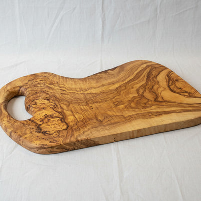 A rustic olive wood serving board with a natural live edge and a cutout handle, displayed on a light wood floor.