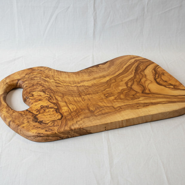 A rustic olive wood serving board with a natural live edge and a cutout handle, displayed on a light wood floor.
