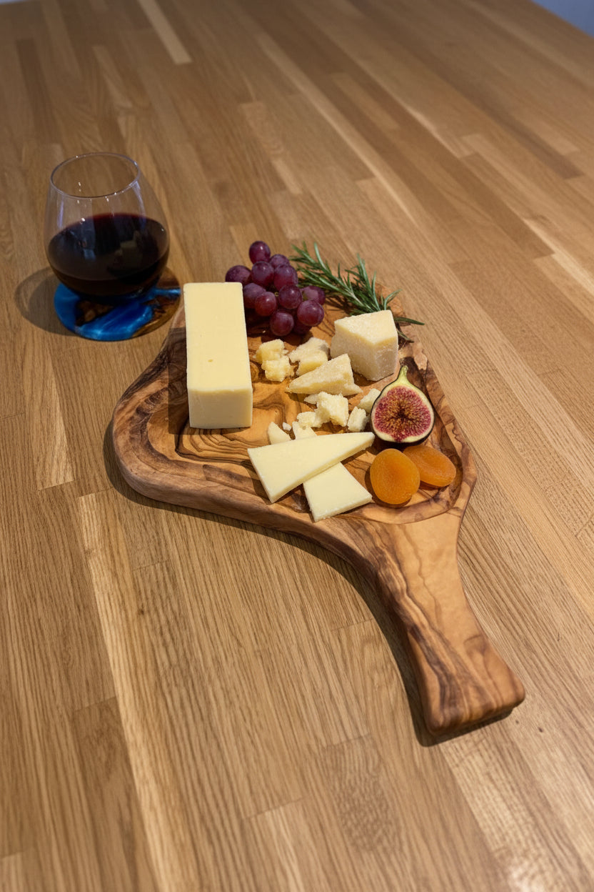 A wooden cutting board with a live edge, shown on a wooden floor with cheese wedges and a large chef's knife. A glass of red wine is visible in the background.