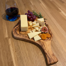 A wooden cutting board with a live edge, shown on a wooden floor with cheese wedges and a large chef's knife. A glass of red wine is visible in the background.