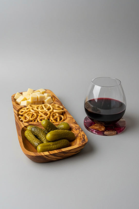 A wooden serving tray with three compartments holds cubed cheese, pretzels, and pickles, placed on a wooden table beside a stemmed wine glass filled with red wine on a coaster.