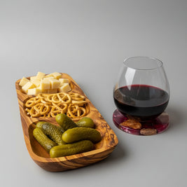 A wooden serving tray with three compartments holds cubed cheese, pretzels, and pickles, placed on a wooden table beside a stemmed wine glass filled with red wine on a coaster.