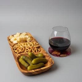 A wooden serving tray with cheese cubes, pretzels, and pickles sits on a wooden table next to a stemmed glass of red wine on a cork coaster.