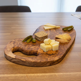 A rustic wooden charcuterie board with cheese wedges and pickle slices, styled on a wooden table with two filled cocktail glasses in a home setting.