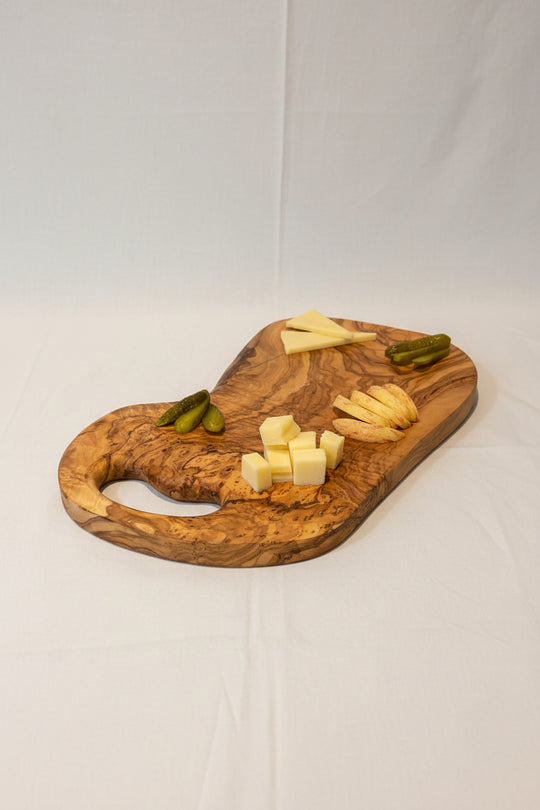 A wooden charcuterie board with cheese wedges and cubes, sliced apples, pickles, and two cocktail glasses on a wooden table in a home setting.