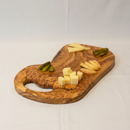 A wooden charcuterie board with cheese wedges and cubes, sliced apples, pickles, and two cocktail glasses on a wooden table in a home setting.