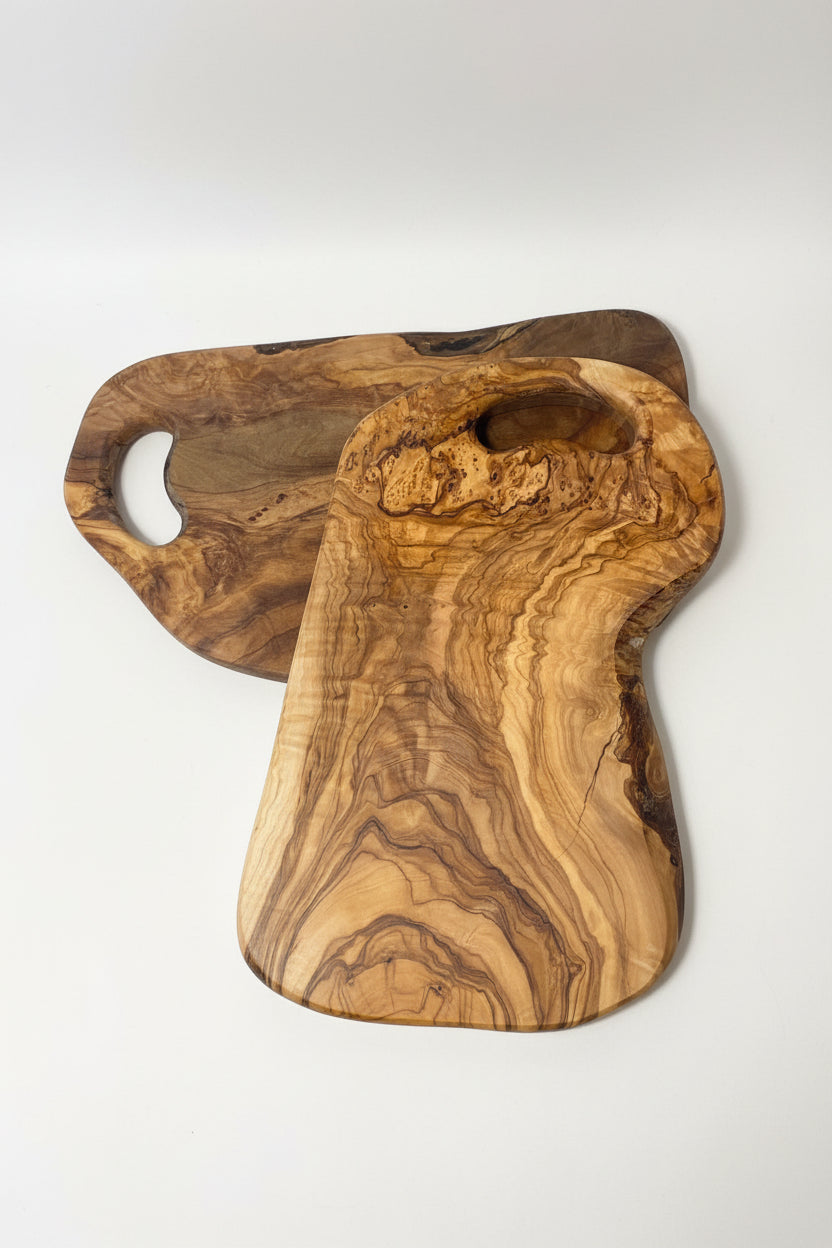 Two handcrafted olive wood cutting boards with natural live edges and grain patterns, displayed on a light wood floor.