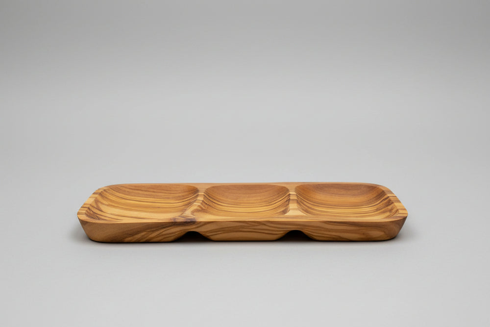 A wooden serving tray with three compartments and a wavy grain pattern, placed on a wooden table next to a glass of red wine.
