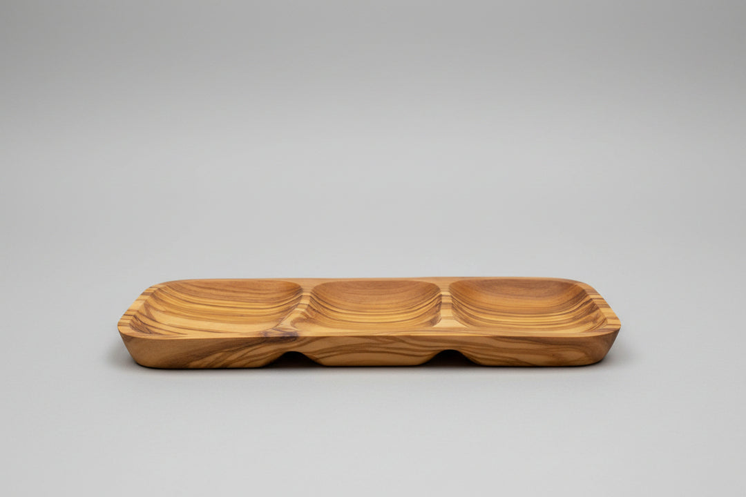 A wooden serving tray with three compartments and a wavy grain pattern, placed on a wooden table next to a glass of red wine.