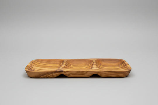 A wooden serving tray with three compartments and a wavy grain pattern, placed on a wooden table next to a glass of red wine.