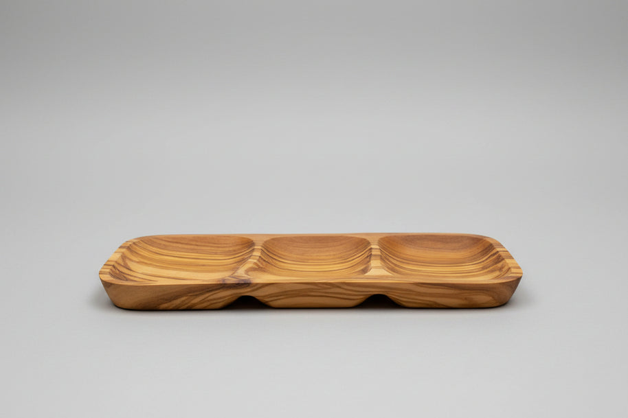 A wooden serving tray with three compartments and a wavy grain pattern, placed on a wooden table next to a glass of red wine.