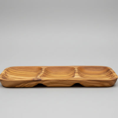 A wooden serving tray with three compartments and a wavy grain pattern, placed on a wooden table next to a glass of red wine.