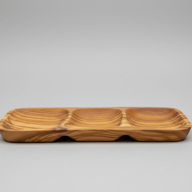 A wooden serving tray with three compartments and a wavy grain pattern, placed on a wooden table next to a glass of red wine.