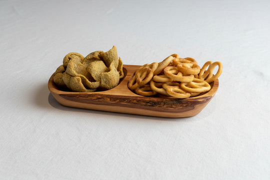 A wooden serving board with a natural bark edge, holding a pile of golden-brown pretzels and a pile of green potato chips, set on a wooden tabletop.