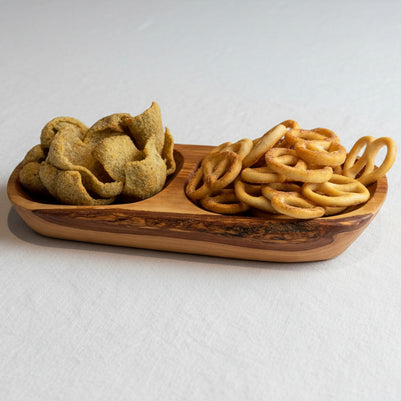 A wooden serving board with a natural bark edge, holding a pile of golden-brown pretzels and a pile of green potato chips, set on a wooden tabletop.