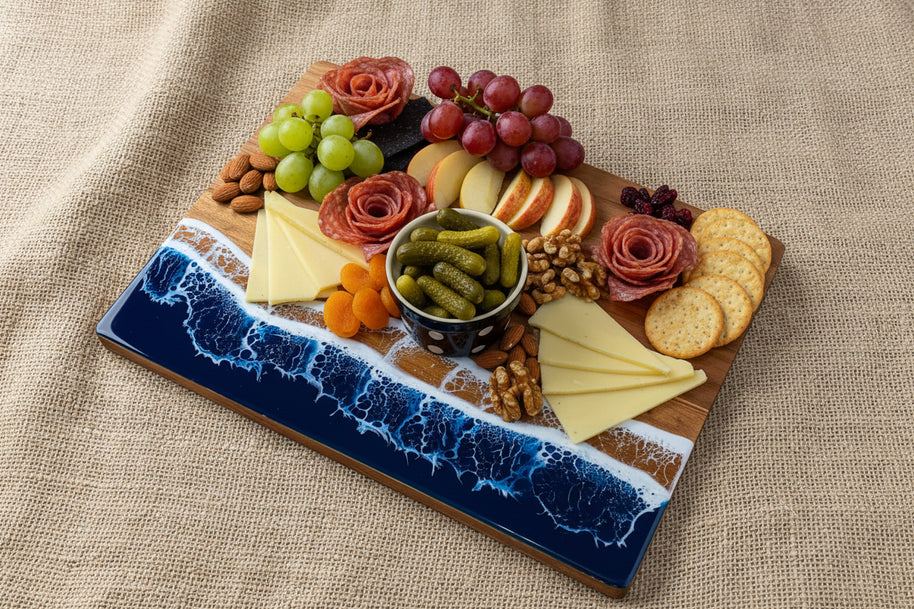 A rectangular wooden charcuterie board with a blue ocean wave design, styled with cheese wedges, crackers, apple slices, and a small bowl of pickles on a burlap surface