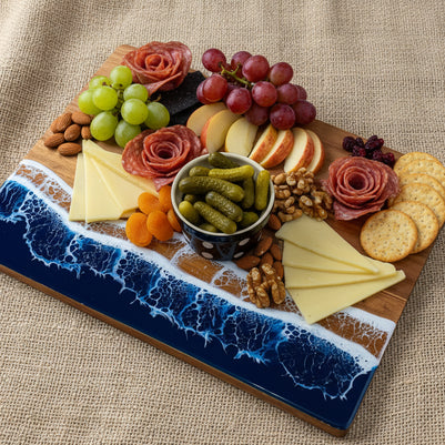 A rectangular wooden charcuterie board with a blue ocean wave design, styled with cheese wedges, crackers, apple slices, and a small bowl of pickles on a burlap surface