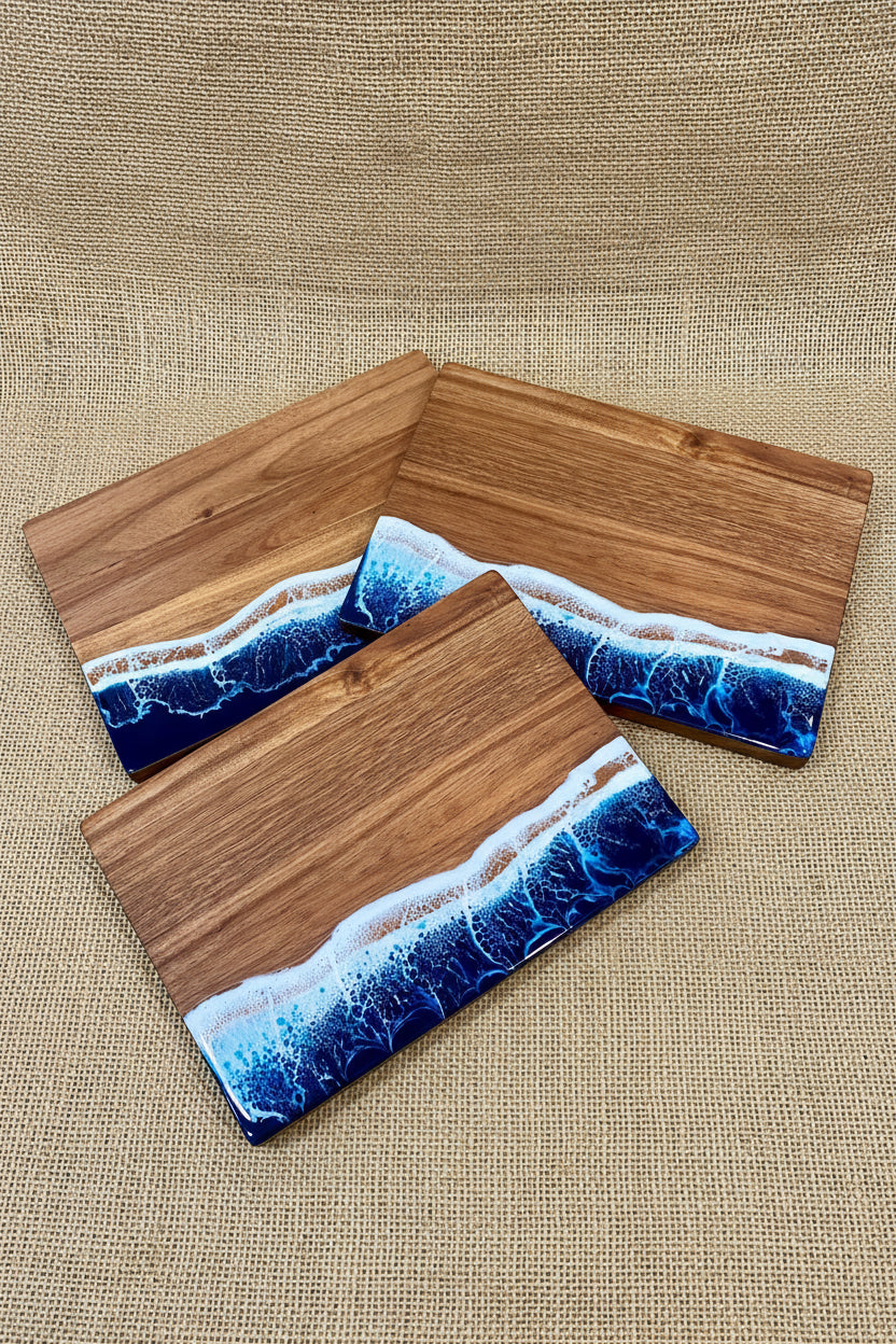 Three rectangular wooden serving boards with a resin ocean wave design in blue, white, and tan, arranged on a burlap surface.