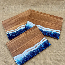 Three rectangular wooden serving boards with a resin ocean wave design in blue, white, and tan, arranged on a burlap surface.