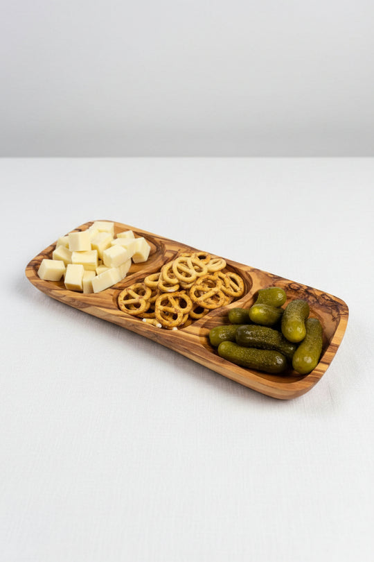A rectangular wooden serving platter with three compartments, filled with cubed cheese, pretzels, and pickles, placed on a wooden tabletop.