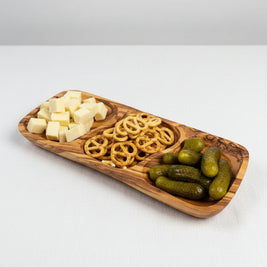 A rectangular wooden serving platter with three compartments, filled with cubed cheese, pretzels, and pickles, placed on a wooden tabletop.