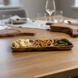 An olive wood three-compartment serving tray holding cubed cheese, pretzels, and pickles on a wooden tabletop.