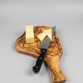 A wooden serving board with a large chef's knife and cheese wedges, placed on a wooden table next to a glass of red wine.