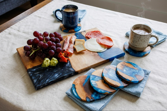 A styled tabletop scene with a wooden and dark blue serving board holding grapes, cheese, salami, and a small green item, alongside two ceramic mugs and matching round coasters on a light fabric-covered table.
