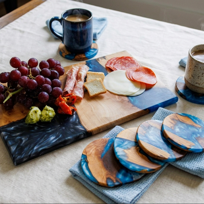 A styled tabletop scene with a wooden and dark blue serving board holding grapes, cheese, salami, and a small green item, alongside two ceramic mugs and matching round coasters on a light fabric-covered table.