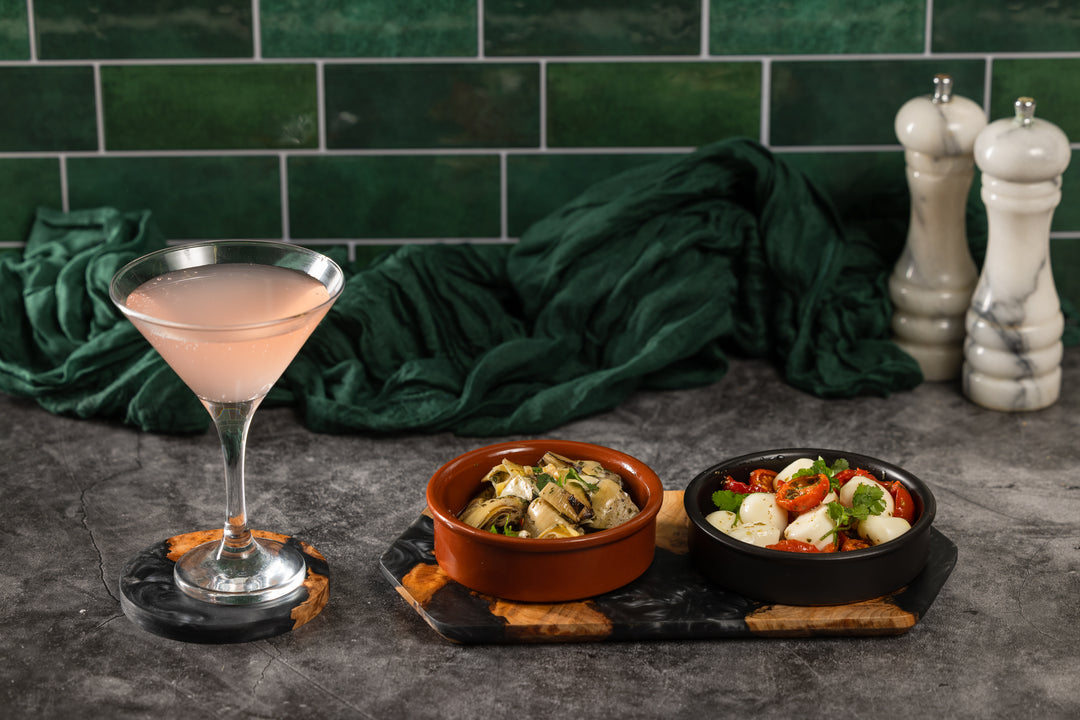 Cocktail glass with a pink drink on a dark surface with two small bowls of food and green tiled wall in the background.