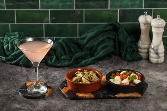 Cocktail glass with a pink drink on a dark surface with two small bowls of food and green tiled wall in the background.