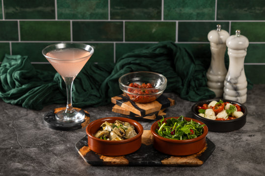 Cocktail glass with a pink drink on a dark surface with bowls of food and a green tiled wall.