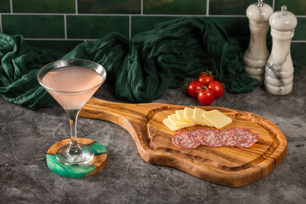 A styled food and drink scene on a dark stone surface: a pink cocktail in a martini glass on a green-and-brown coaster, a wooden serving board with cheese slices, salami, tomatoes, dark green fabric, and two white pepper mills in the background.