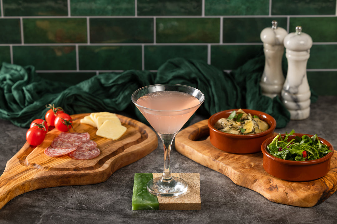 A pink cocktail in a martini glass is styled on a dark stone surface with charcuterie boards, sliced salami, cheese, cherry tomatoes, and two small bowls of salad, set against a green tiled wall and dark green cloth backdrop.