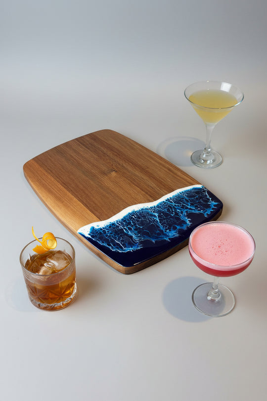 Wooden cutting board with a blue resin design, accompanied by three cocktails on a white surface.