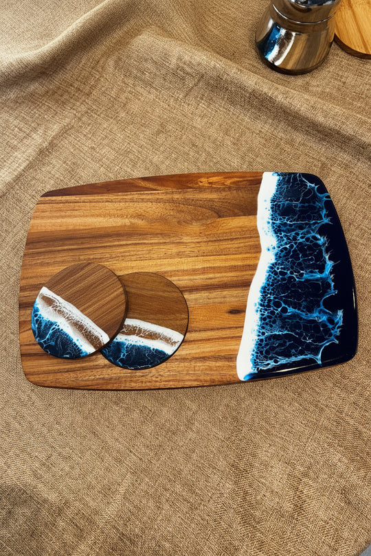 A wooden serving board with a large rounded rectangular section and two oval coasters, decorated with a dark blue and white wave resin design, displayed on a textured beige fabric background.