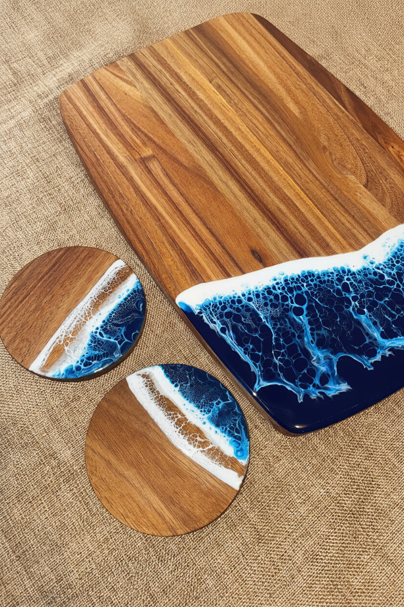 A wooden serving board and two matching coasters with a blue ocean wave and white foam design, arranged on a textured beige fabric background.