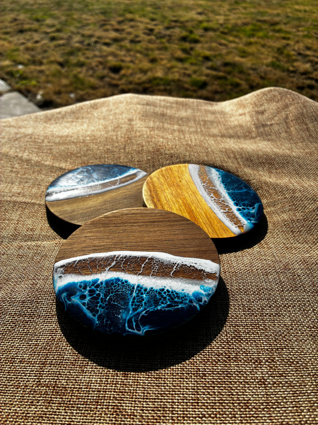 Three round coasters with a resin ocean wave design resting on a brown burlap fabric outdoors.
