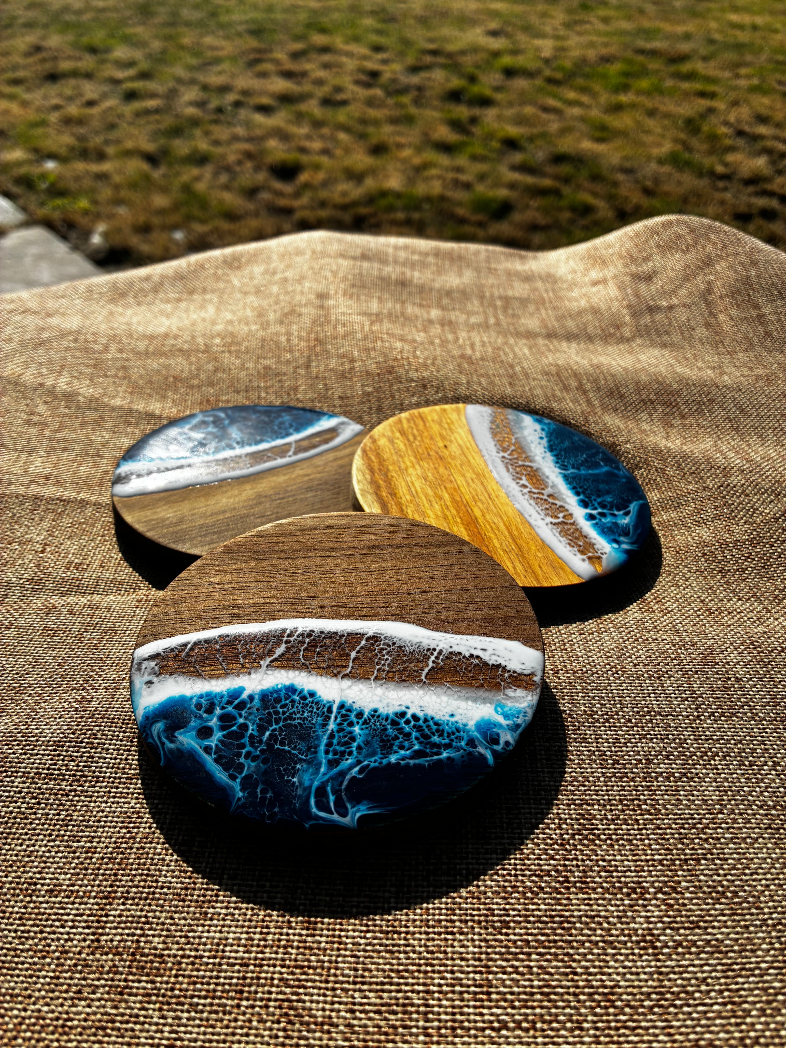 Three round coasters with a resin ocean wave design resting on a brown burlap fabric outdoors.