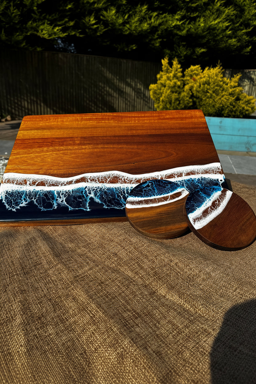 A rectangular wooden serving board with a heart-shaped wooden top, featuring a wave-like resin inlay in shades of blue, white, and dark gray, photographed outdoors on a woven mat with trees in the background.