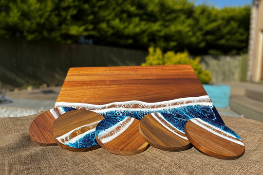 A rectangular wooden serving board with a wave design in blue, white, and brown resin, accompanied by four matching round coasters, displayed outdoors on a burlap surface with a blurred garden background.