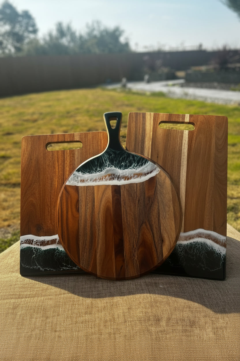 A round wooden serving board with a handle, displayed outdoors on a burlap surface with a matching rectangular wooden board behind it. The boards feature a natural brown wood grain base with a resin inlay depicting ocean waves in white and dark gray.