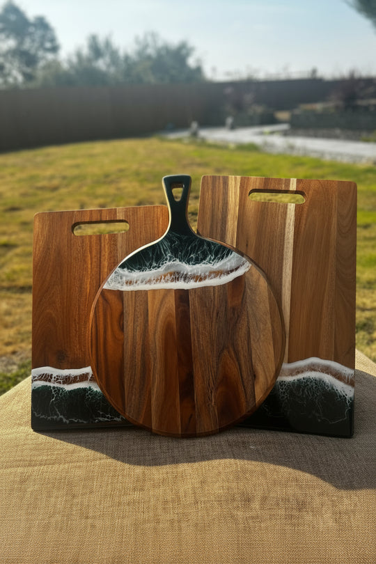 A round wooden serving board with a handle, displayed outdoors on a burlap surface with a matching rectangular wooden board behind it. The boards feature a natural brown wood grain base with a resin inlay depicting ocean waves in white and dark gray.