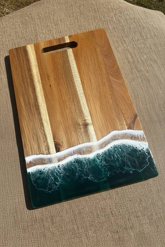 A rectangular wooden cutting board with a handle cutout, featuring a printed or inlaid ocean wave design in dark teal and white foam along one side, placed on a textured brown fabric surface.